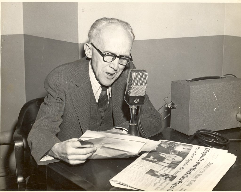 Black and white photo of newsman Al Sigl holding a newspaper and speaking into a microphone as part of his daily radio broadcast