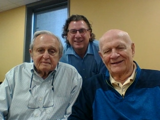 Two men seated at a table facing the camera with one man standing behind them; all are smiling