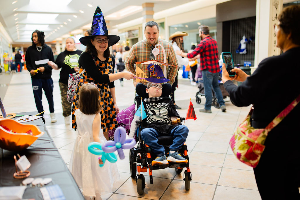 a group is taking a photo together; one adult is wearing a witch hat; the boy in the center uses a wheelchair and is also wearing a costume