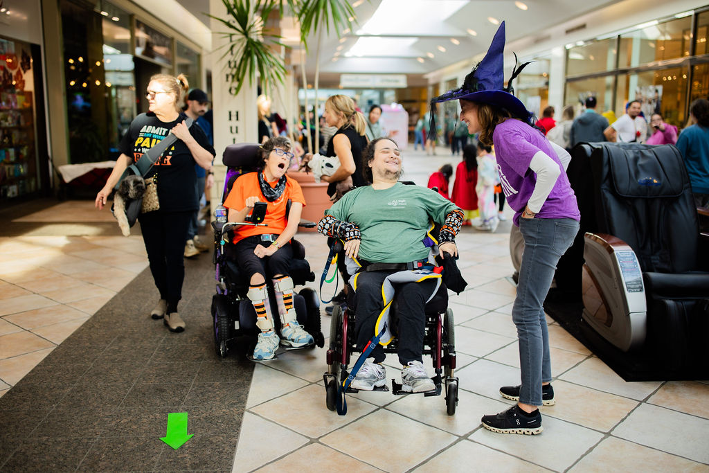 two women who use wheelchairs smiling and talking with a woman wearing a witch hat costume