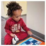 a young boy with curly brown hair wearing a red outfit sitting on the floor ready to choose a word on his communication device