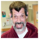 a man with dark hair and a salt and pepper beard wearing a dark red hoodie, smiling into the camera