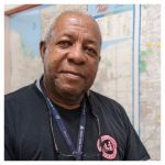 an older man wearing a black shirt standing in front of a large map of Rochester smiling