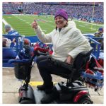 a smiling woman wearing a Buffalo Bills knit cap and white coat giving a thumbs-up riding a motorized scooter at a football game