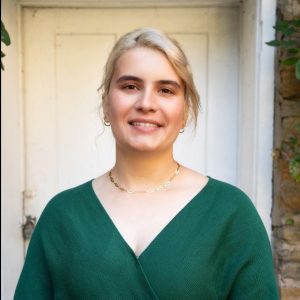 a woman smiling into the camera, her blonde hair is pulled back and she is wearing a green top; a doorway is visible behind her