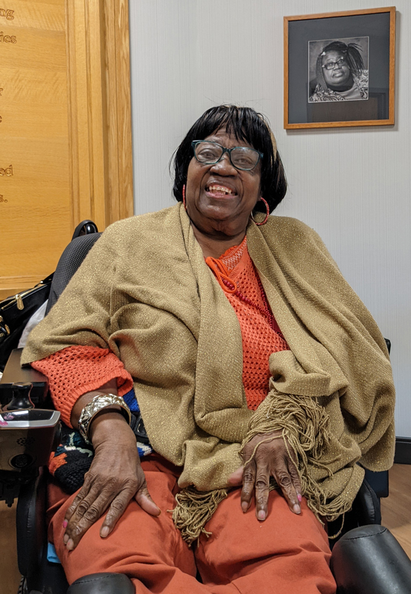 an older woman wearing glasses and a tan shawl who uses a wheelchair; she is indoors in front of her framed portrait on the wall