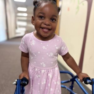 a young girl smiles into the camera wearing a light purple dress and using a blue pediatric stander