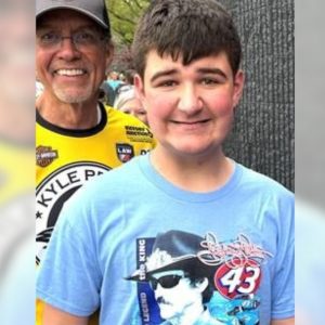 a teen with short dark hair wearing a blue NASCAR tshirt smiles into the camera with another man wearing a baseball cap and yellow shirt stands smiling in the background