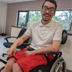 a young man with short dark hair and glasses smiles happily into the camera wearing a white shirt and red pants and he uses a wheelchair