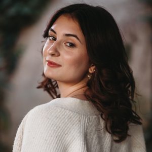 a young woman with shoulder length dark hair looks back over her shoulder into the camera wearing a white sweater