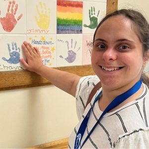 a woman with dark hair smiles into the camera; she is wearing a white shirt with blue stripes and is holding her hand up to a much smaller handprint with her name below it in blue paint that is part of a mural on the wall