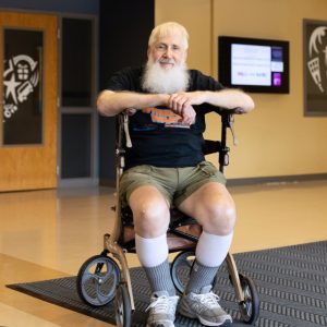 a man with gray hair and beard looks into the camera wearing a black tshirt that says Wheels for Warren and he is seated in a rolling walker