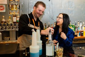 a man and a woman laugh together making drinks behind a bar