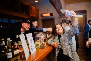 a woman taking a selfie with a man smiling leaning into the frame from behind the bard
