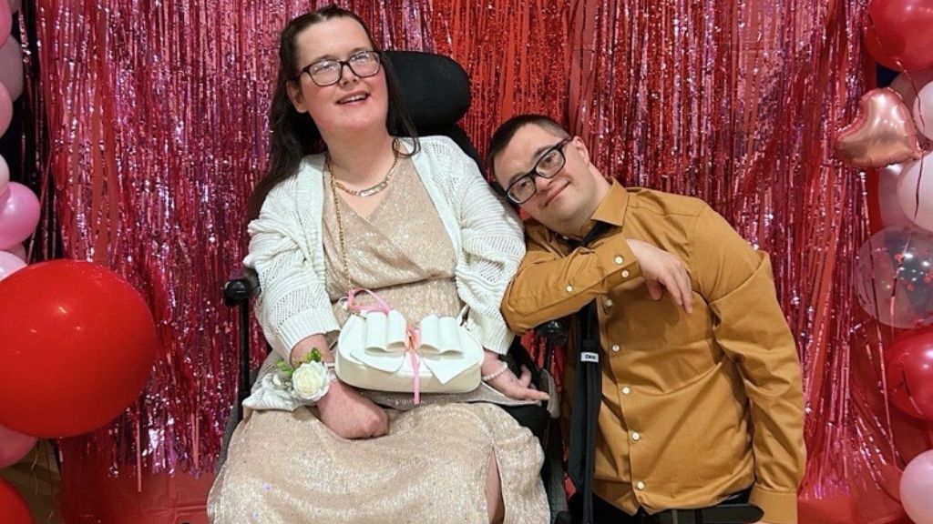 a young woman with long brown hair wearing a long dress; a young man leans his head on her shoulder; she is a wheelchair user