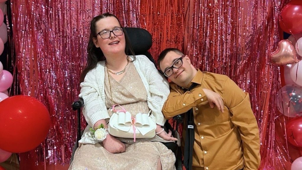a young woman with long brown hair wearing a long dress; a young man leans his head on her shoulder; she is a wheelchair user